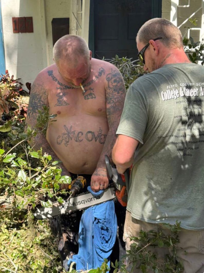 Two shirtless men clear storm debris outside a house; one, heavily tattooed with GAME OVER across his stomach, cigarette in his mouth, holds a drill while the other steadies a chainsaw.